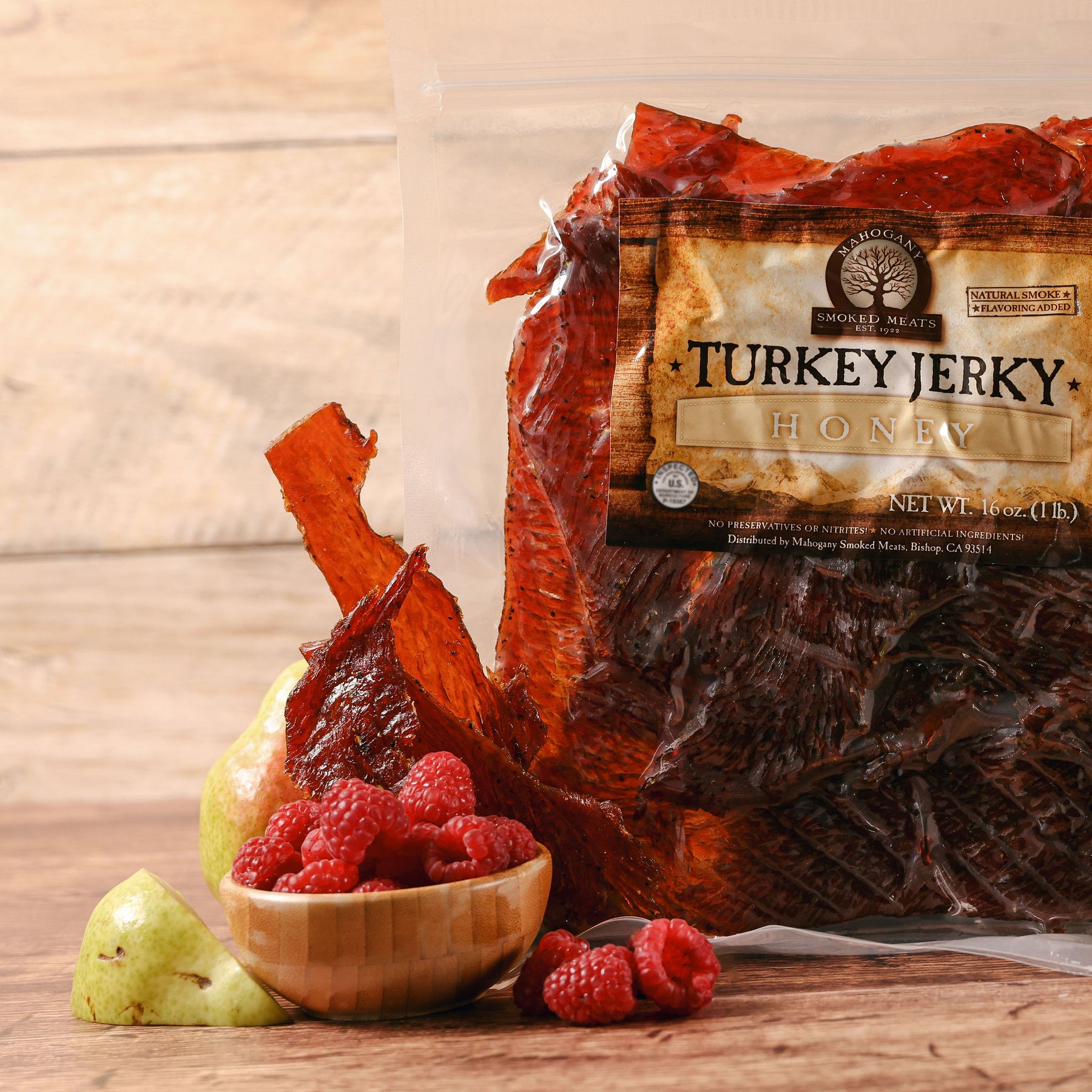 Bag of honey turkey jerky with slices visible, accompanied by raspberries and a pear on a wooden surface.
