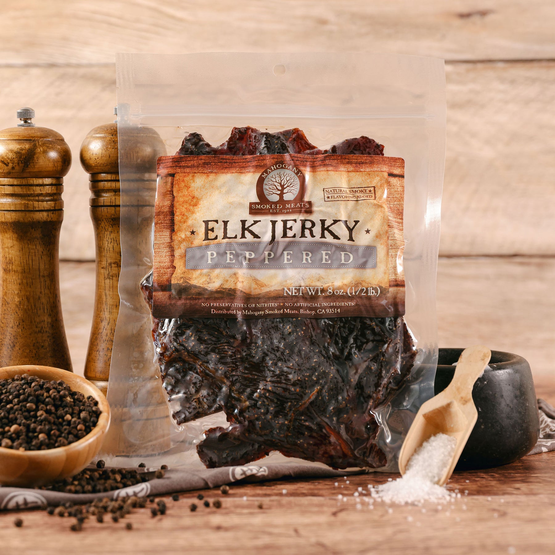 Package of peppered elk jerky displayed with a mortar of black peppercorns and salt, wooden shakers at back.
