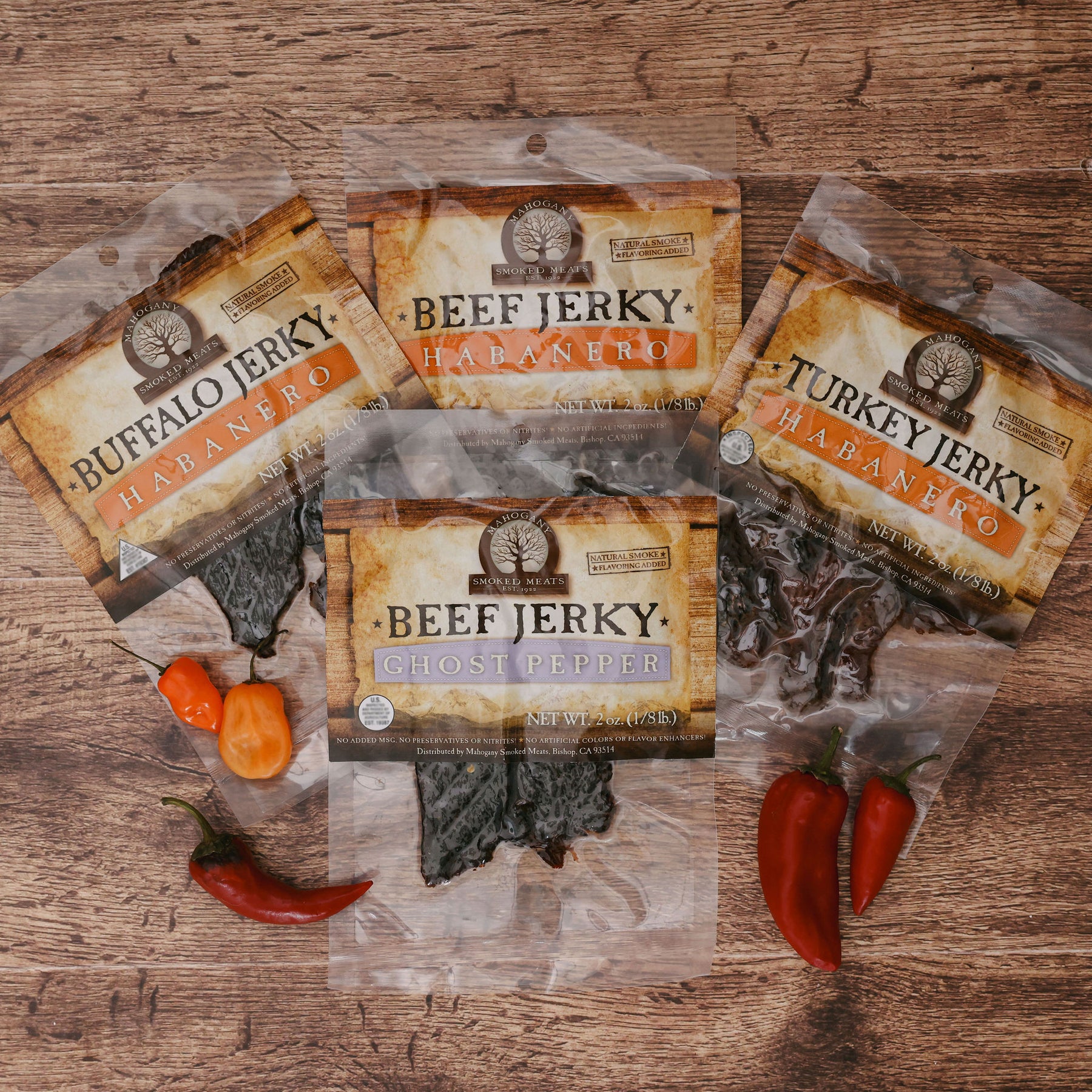 Four varieties of spicy jerky packages on a wooden surface with habanero and ghost peppers