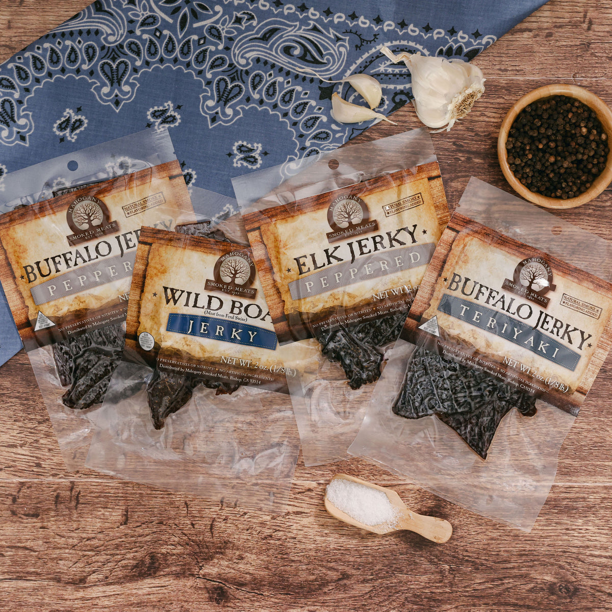 Variety of exotic jerky packets (buffalo, elk) displayed on wooden surface with spices and bandana.