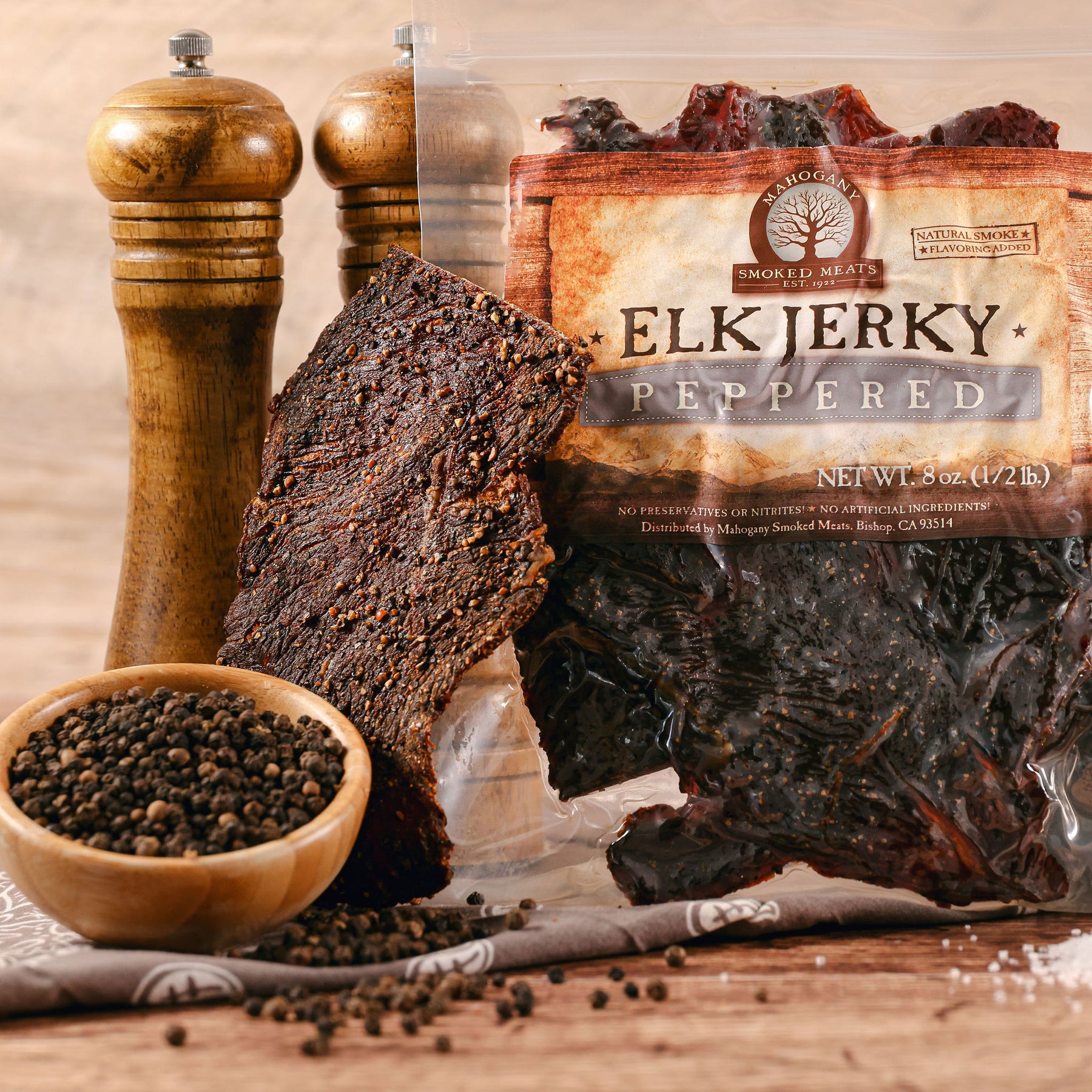 Elk jerky with black pepper, wooden pepper grinders, and peppercorns on a rustic table.