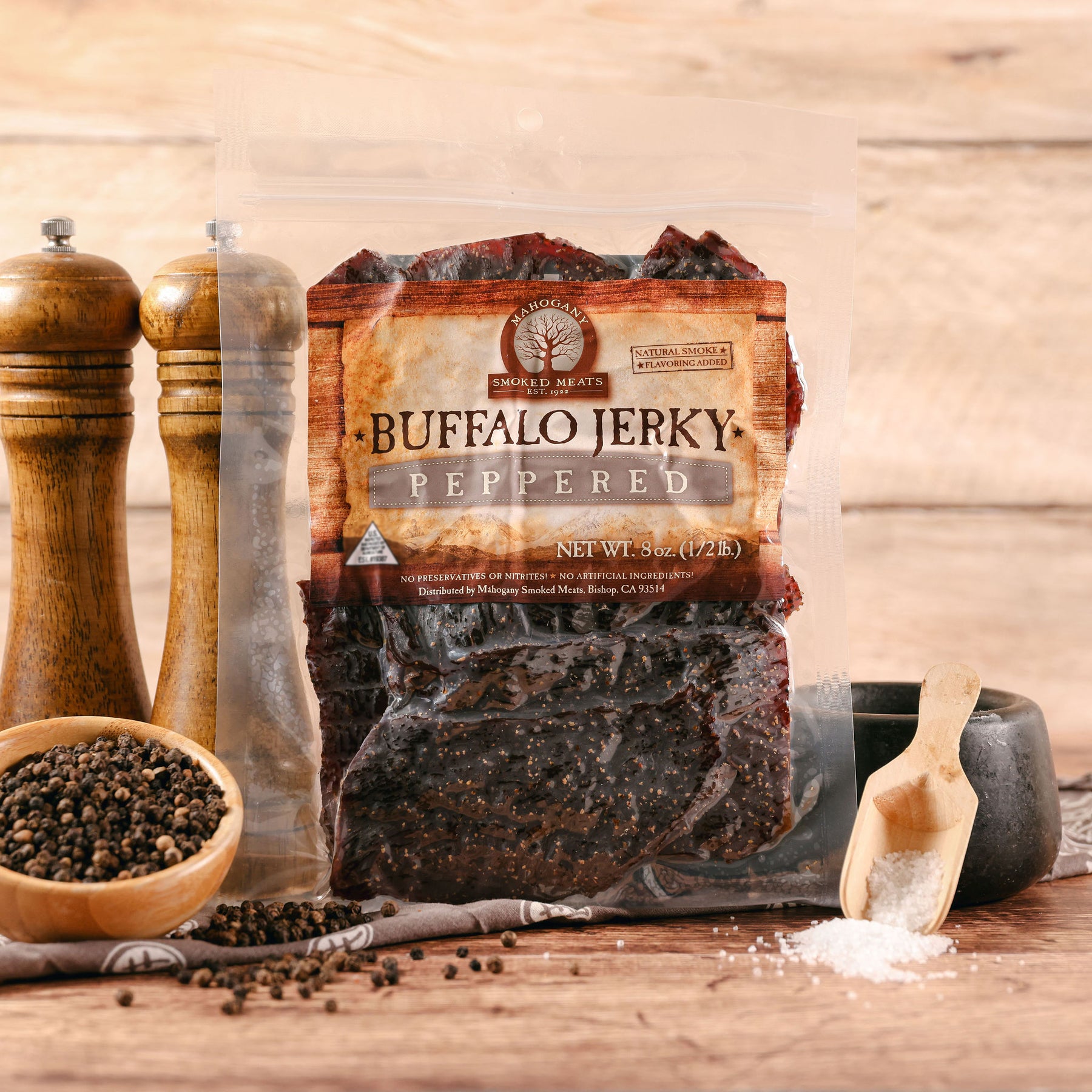 Packaged buffalo jerky with pepper, flanked by pepper mills and scattering of whole peppercorns on wooden surface.