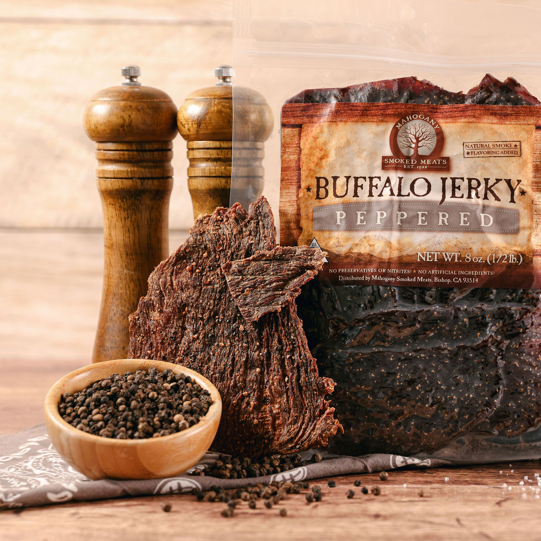 Buffalo jerky pieces with black pepper, salt grinders, and peppercorns on a wooden table