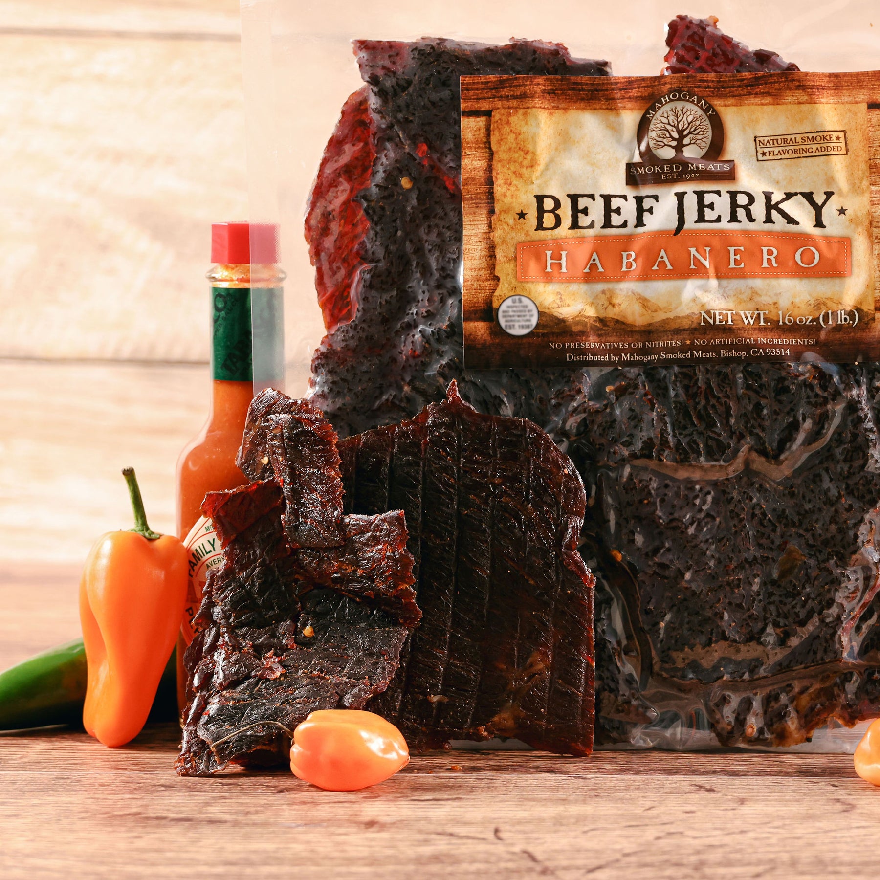 Beef jerky pieces and habanero peppers beside a bottle of hot sauce on a wooden surface.