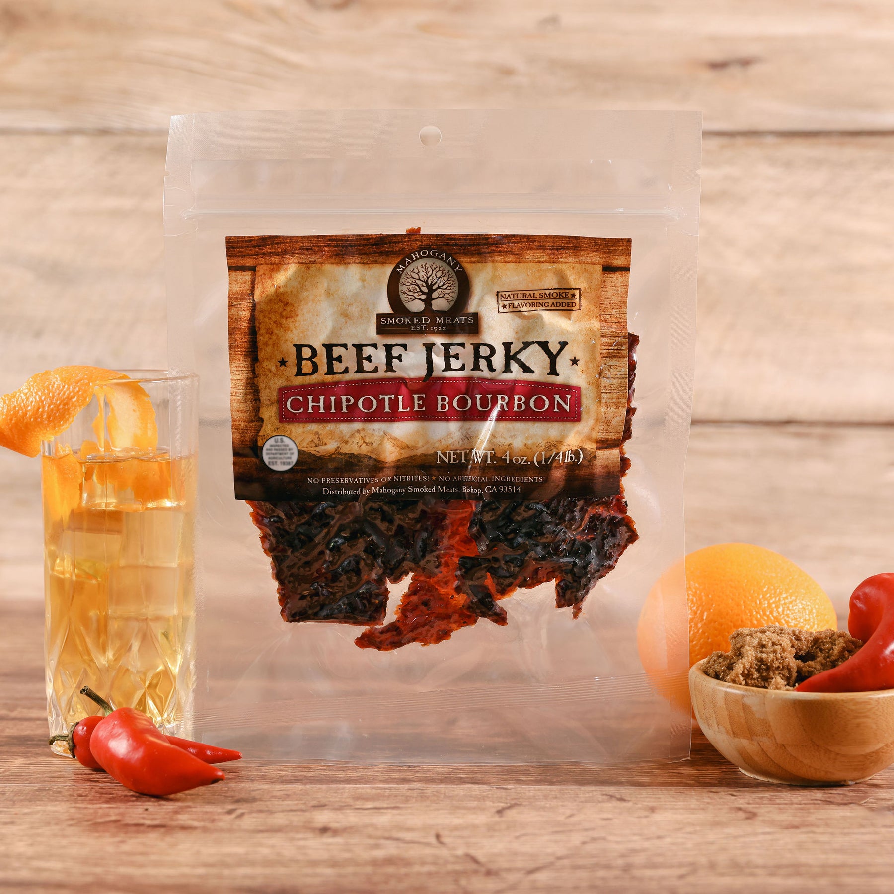 Packaged chipotle bourbon beef jerky displayed with an orange garnished drink, red chilis, and brown sugar.