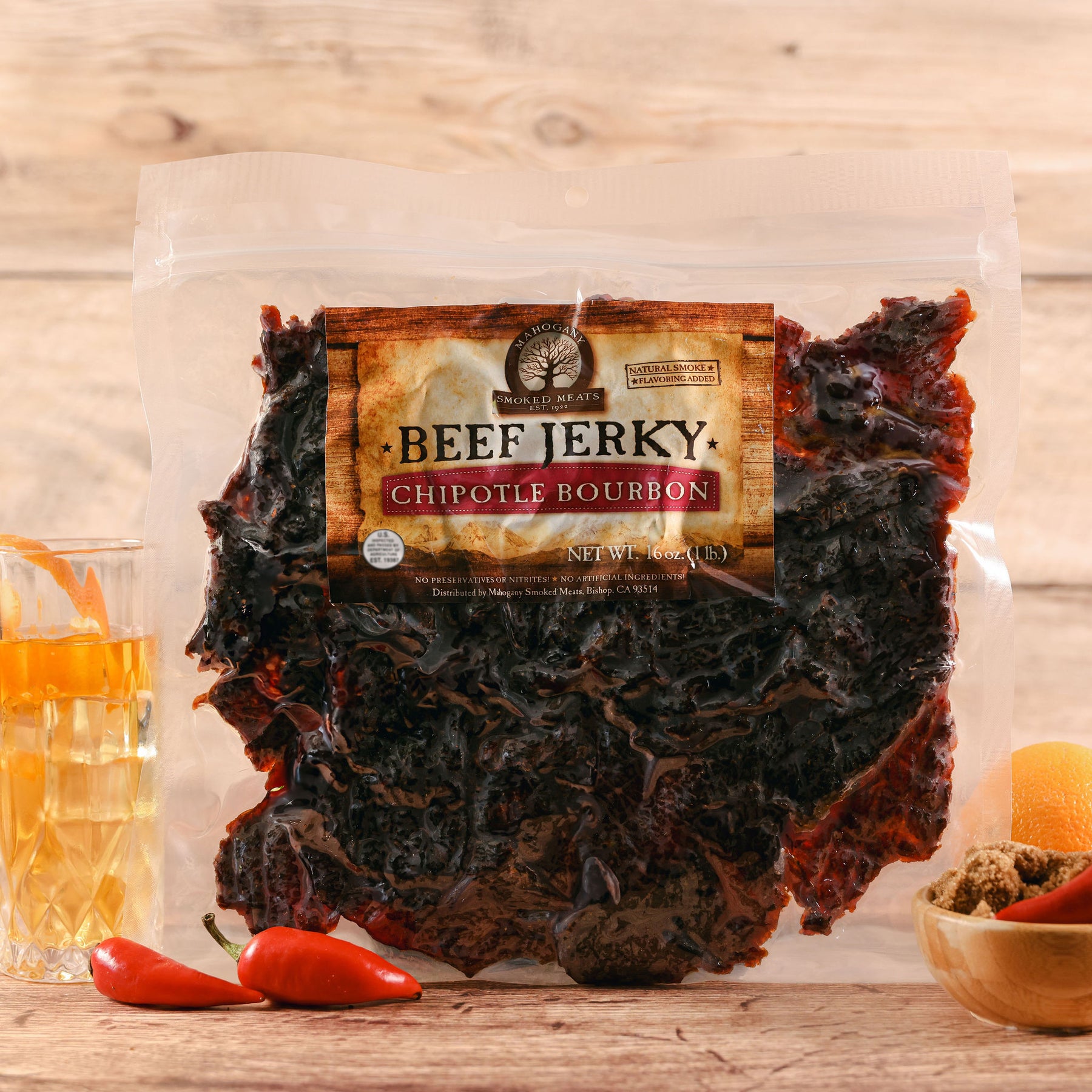 Packaged chipotle bourbon beef jerky with visible slices, accompanied by red chili peppers and a glass of bourbon.