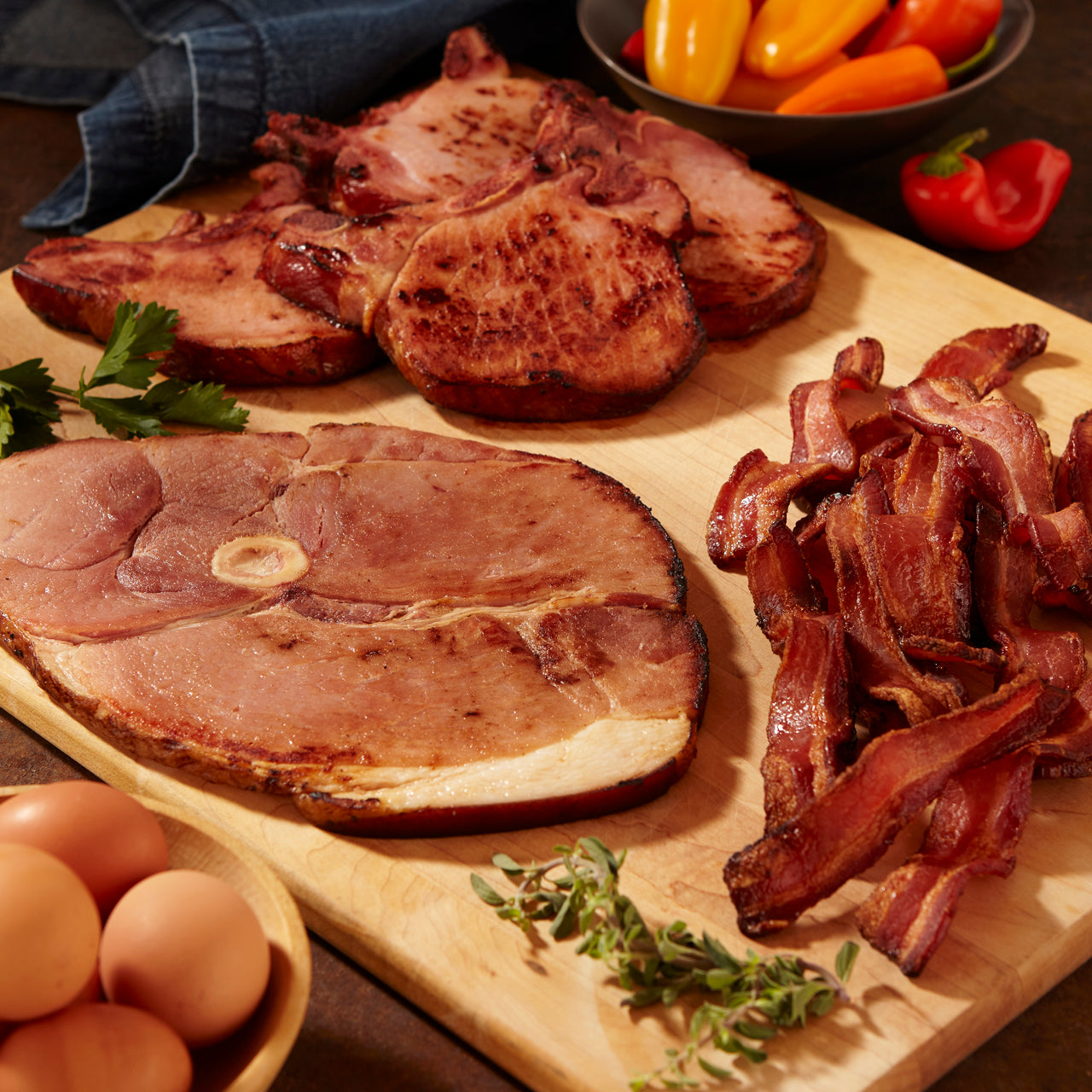 Assorted cooked meats including ham slices and crispy bacon on a wooden board, with a bowl of peppers in the background.