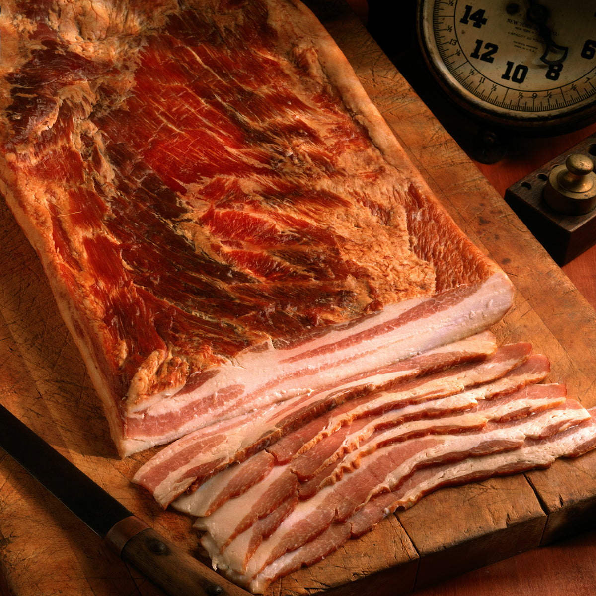 Half slab of bacon on a wooden board with slices cut alongside an antique scale.