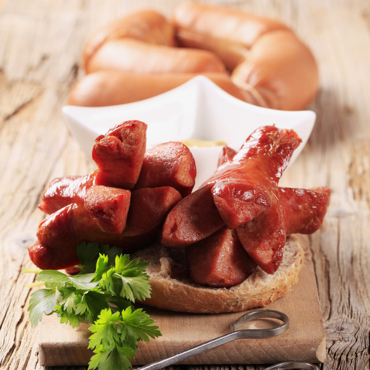 Sliced knackwurst on bread with whole sausages in background, garnished with parsley on wooden table.