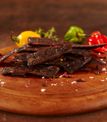 Habanero brisket jerky on a wooden board with colorful peppers and scattered spices.