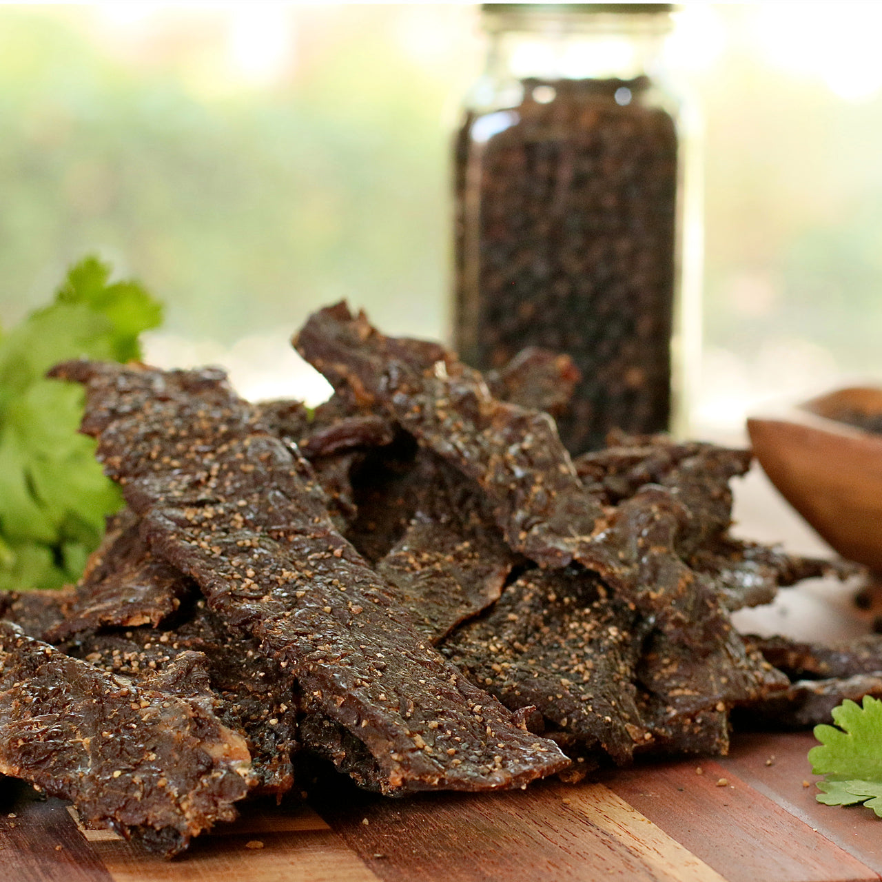 Peppered brisket jerky pieces on a wooden board with a jar of peppercorns in the background