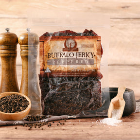 Packaged buffalo jerky with pepper, flanked by pepper mills and scattering of whole peppercorns on wooden surface.