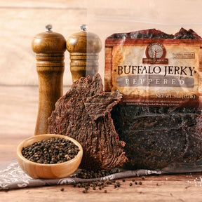 Buffalo jerky pieces with black pepper, salt grinders, and peppercorns on a wooden table