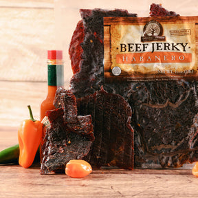 Beef jerky pieces and habanero peppers beside a bottle of hot sauce on a wooden surface.