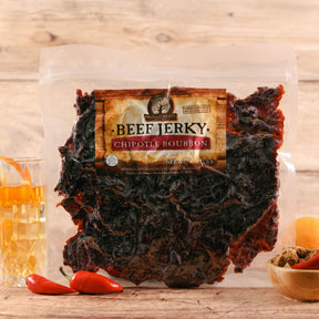 Packaged chipotle bourbon beef jerky with visible slices, accompanied by red chili peppers and a glass of bourbon.