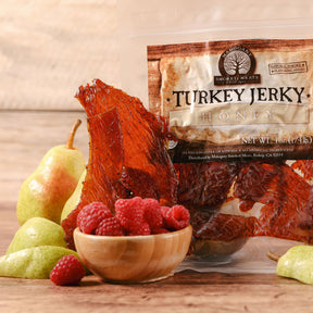 Glistening honey turkey jerky displayed beside a bowl of raspberries and pears on a wooden surface.