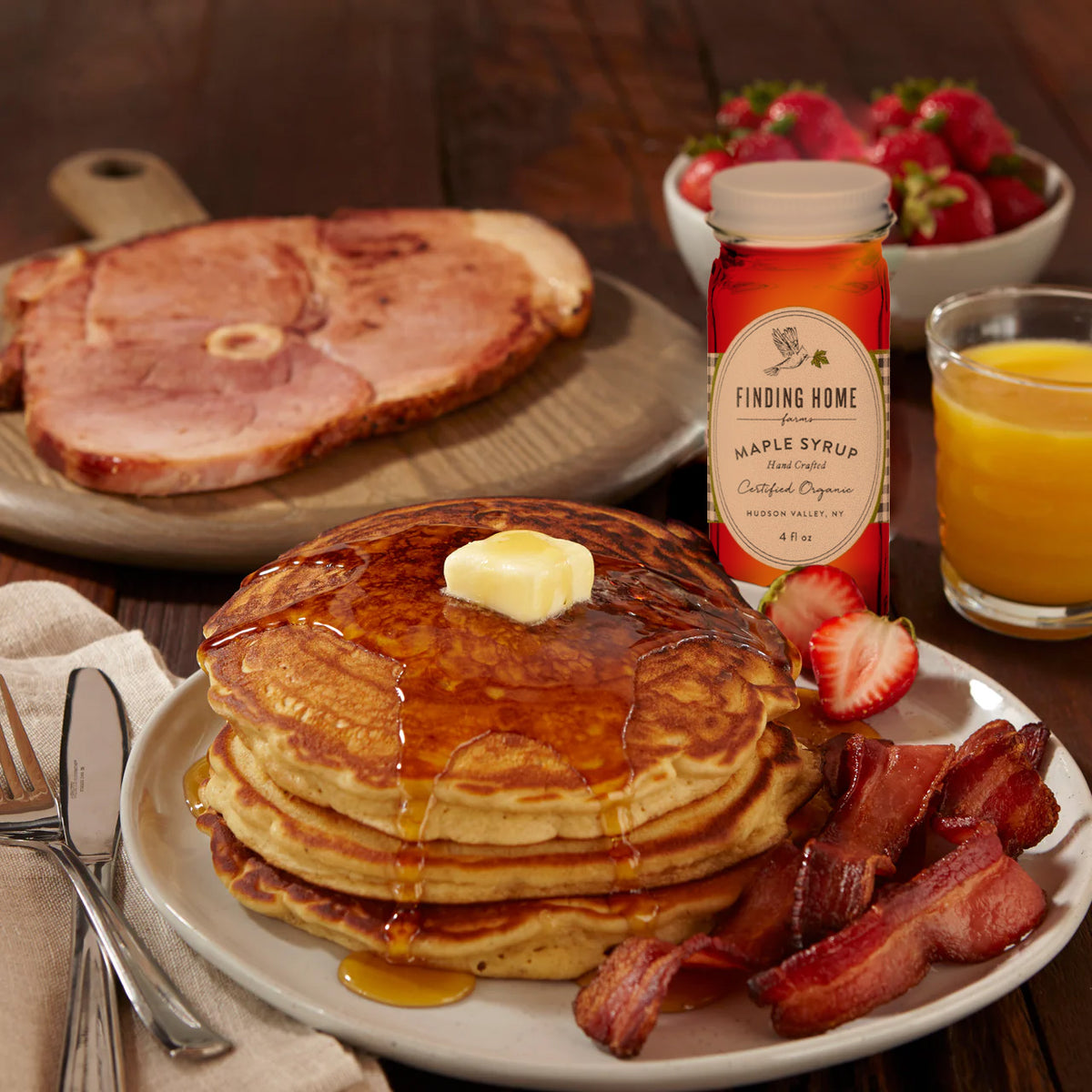A breakfast layout showing pancakes, smoked bacon, ham steak, maple syrup and orange juice.