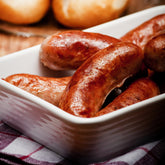 Grilled breakfast sausages in a white dish with blurred dinner rolls in background.